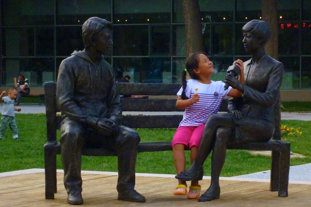 A small child sits on a bench next to a statue of a female broadcast reporter, smiles, and puts a hand on her microphone. Opposite the reporter is a statue of a male interviewee, who is not saying much because he is a statue.